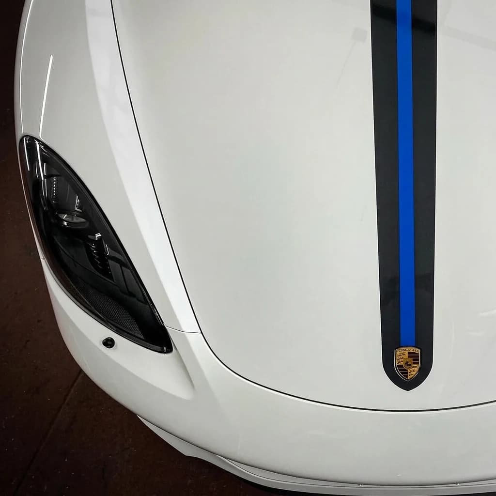 Plan serré bas sur le capot d'une Porsche 718 Cayman T blanche, bande bleue centrale, écusson Porsche et optique avant