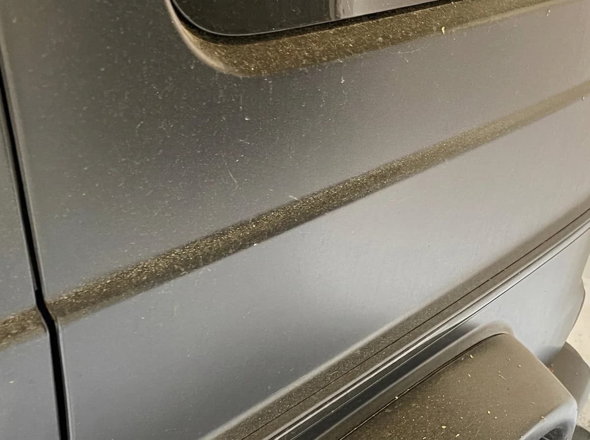 Plan serré sur la porte arrière et le marchepied d'une Mercedes-AMG G63 noir mat, couverts de poussière avant lavage
