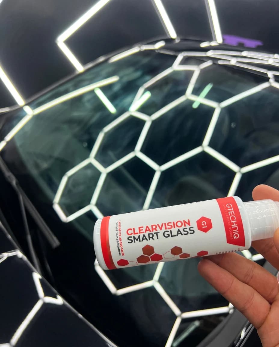 Flacon de traitement vitrage GTechniq ClearVision tenu en main devant un pare-brise noir, sous les LEDs hexagonales