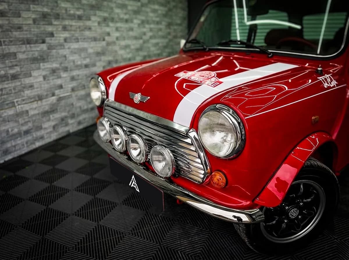 Plan serré sur la calandre chromée d'une Mini classique rouge, ses bandes blanches et ses quatre phares longue portée