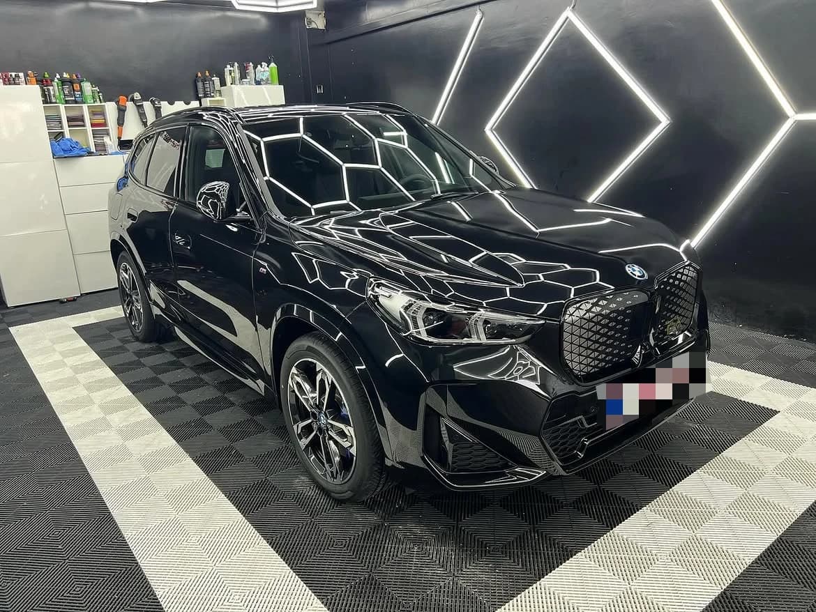 BMW iX1 noire de trois quarts avant en plan large dans l'atelier ISA, avec les lignes des LEDs réfléchies sur la carrosserie