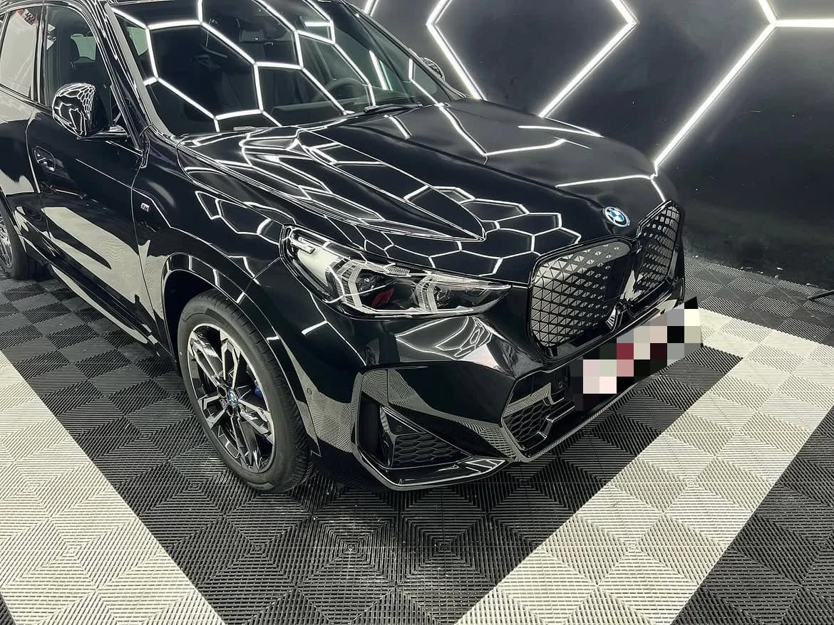 Plan serré sur une BMW iX1 noire de trois quarts avant dans l'atelier, la calandre et le capot traversés par les reflets des LEDs