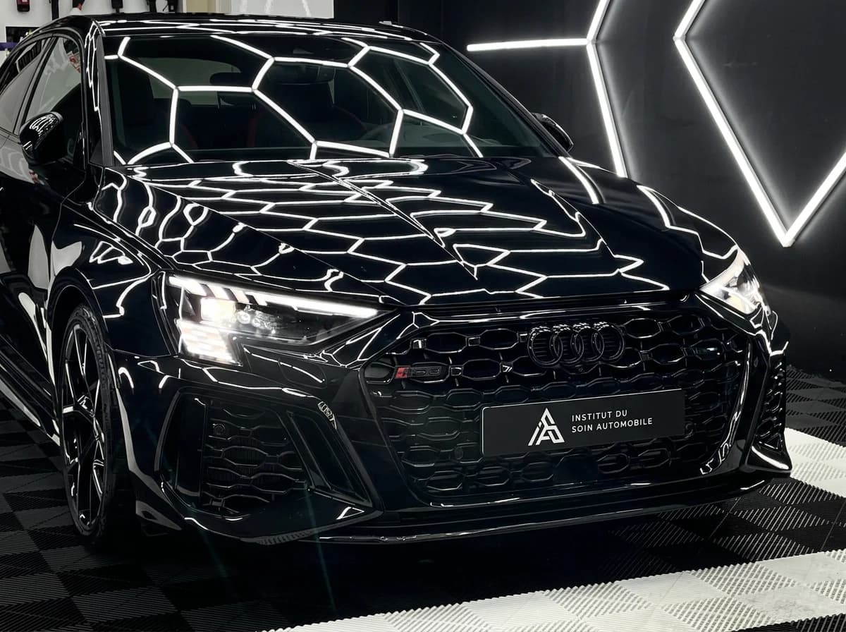Audi RS3 noire de trois quarts avant dans le studio de detailing, les LEDs hexagonales du plafond réfléchies sur la carrosserie